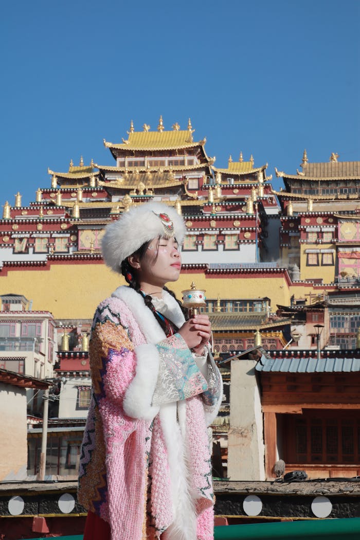 A woman in traditional Tibetan attire with Ganden Sumtsenling Monastery in the background, under a clear blue sky.